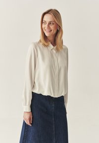 Bluză cu buton, de culoare bej deschis, cu mânecă lungă și guler clasic, asortată cu o fustă A-line din denim de culoare închisă. Material neted, croială lejeră.