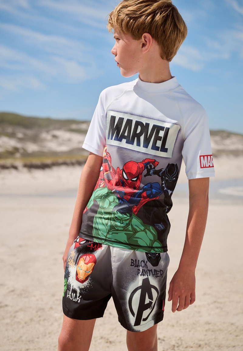 Maglia a maniche corte bianca con logo Marvel e grafiche di supereroi. Pantaloncini neri con vari design di personaggi e colori vivaci.