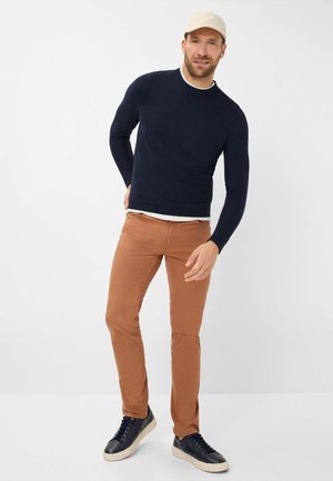 Marineblauer Strickpullover, enganliegend, runder Ausschnitt; braune enge Chinohosen; schwarze Sneakers mit weißen Sohlen; hellfarbige Kappe seitlich getragen.