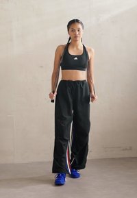 Svart athletic crop top med Adidas-logo, løse svarte bukser med side-striper i rødt, hvitt og blått, og blå sportsko.