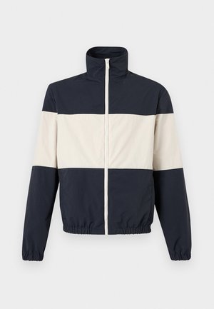 Mladi muškarac stoji sa rukama u džepovima, nosi plavu i bijelu jaknu s blokovima boja, krem džemper, široke bijele hlače i smeđe tenisice.