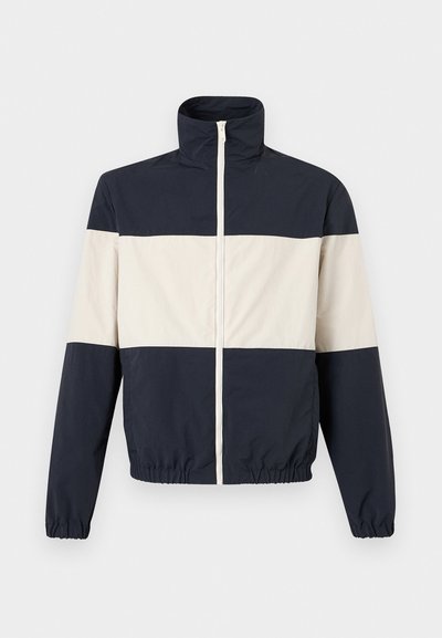 Μπουφάν σε μπλε ναυτικό και λευκό χρώμα με ψηλό γιακά, μπροστινό φερμουάρ, ελαστικά μανίκια και ρίγα.