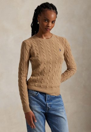 Kobieta z warkoczami, ubrana w beżowy sweter z warkoczowym splotem i niebieskie dżinsy, stojąca na neutralnym tle.