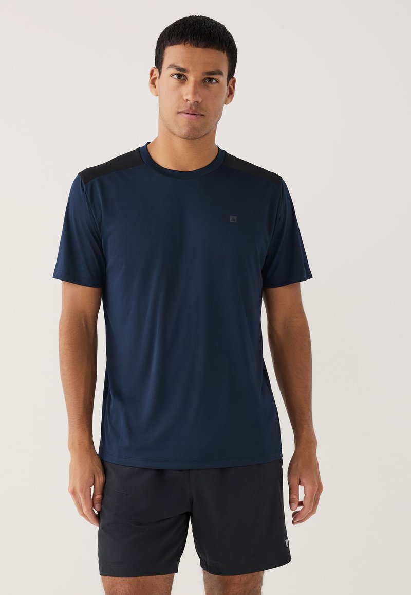 Giovane uomo con una camicia blu navy a maniche corte con spalle nere e pantaloncini sportivi neri, in piedi davanti a uno sfondo semplice.