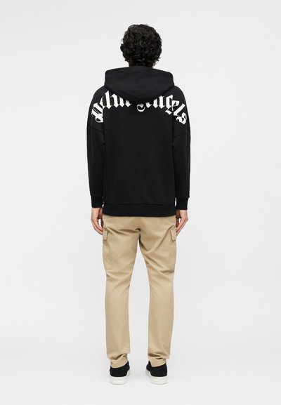 Inimene lokkis tumedate juustega, kes kannab musta kapuutsiga pusa valge "Palm Angels" tekstiga seljal, beeže värvi töörõivaid pükse ja musti tossusid.