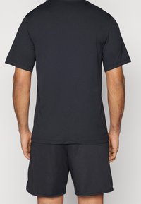 Man som bär en enkel svart kortärmad t-shirt och svarta shorts, sedd bakifrån mot en enkel ljus bakgrund.