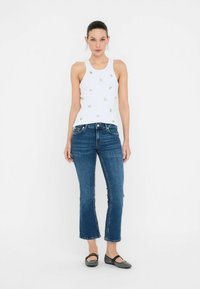 Model staat tegen een effen achtergrond, gekleed in een witte tanktop met decoratieve elementen en blauwe jeans, aangevuld met zwarte ballerina's.