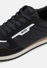Black athletic sneaker with suede and mesh materials, white accents, logo on upper, textured laces, and a light-colored sole with tread.
