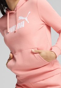 Sudadera con capucha de color rosa, fabricada en un tejido suave, que presenta un bolsillo canguro y un gran logo blanco de Puma en el pecho.