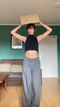 Model wearing a black sleeveless crop top and gray wide-leg trousers, holding a Zalando box above her head against a green wall.