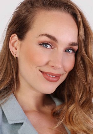 Goldene Kreolen- und Steckerohrringe an einem Modell mit welligem, hellbraunem Haar. Sanfte Make-up-Highlights und ein kragenblauer Blazer.