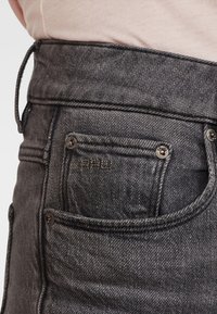 Jeans de mezclilla grises de tiro alto con un acabado texturizado, que presentan un pequeño bolsillo trasero y herrajes de botón de metal. Se pueden ver sutiles detalles de costura.