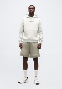 Νεαρός άνδρας στέκεται φορώντας ανοιχτό γκρι φούτερ με κουκούλα, μπεζ σορτς μέχρι το γόνατο, λευκές κάλτσες και λευκά αθλητικά παπούτσια Nike, μπροστά σε απλό φόντο.