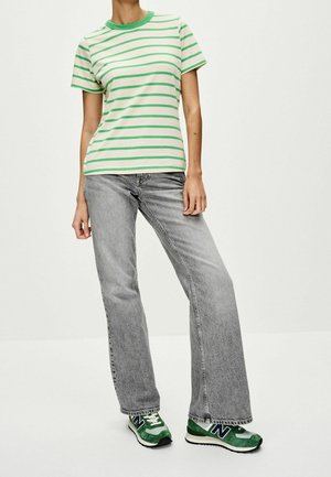 Osoba ubrana w zielono-białą koszulkę w paski, szare spodnie o szerokich nogawkach oraz zielono-białe sneakersy New Balance, stojąca na jednolitym tle.