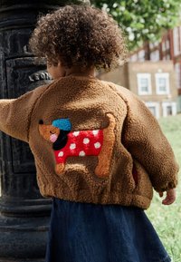 Brun blød fleecejakke med broderet hundedesign i rødt og med prikker, iført en blå hue og med krøllet struktur. Denimnederdel synlig.