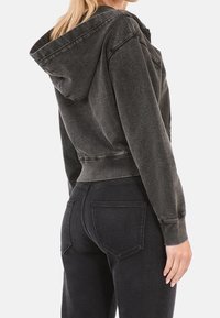 Donkergrijze cropped hoodie van zachte stof, voorzien van een capuchon, geribbelde manchetten en een gladde textuur, gecombineerd met zwarte jeans met achterzakken.