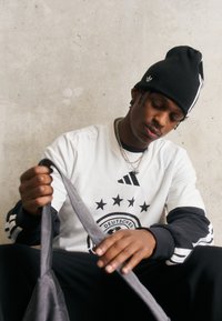 Hvit langermet skjorte med svarte detaljer som har en logo og stjerner; svart strikket lue; gråt stoff som holdes; sitter mot en betongvegg.