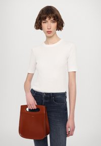 Hnedá kožená taška so strihanými uchami, držaná v ruke. Biely krátky rukáv a tmavé riflové džínsy dopĺňajú outfit.