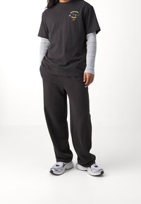 Osoba v černých teplácích Puma, černém tričku s barevným potiskem "Mládež je budoucnost", bílém dlouhém rukávu a bílých teniskách.