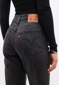 Άτομο που φοράει μακρυμάνικη μαύρη μπλούζα και ψηλό μεσο μαύρο τζιν Levi's, δείχνοντας την πίσω τσέπη και την ετικέτα της μάρκας στη μέση.