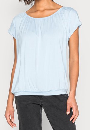 Femme portant un chemisier bleu clair à manches courtes avec un col et un ourlet froncés, associée à un pantalon noir, debout devant un fond uni.