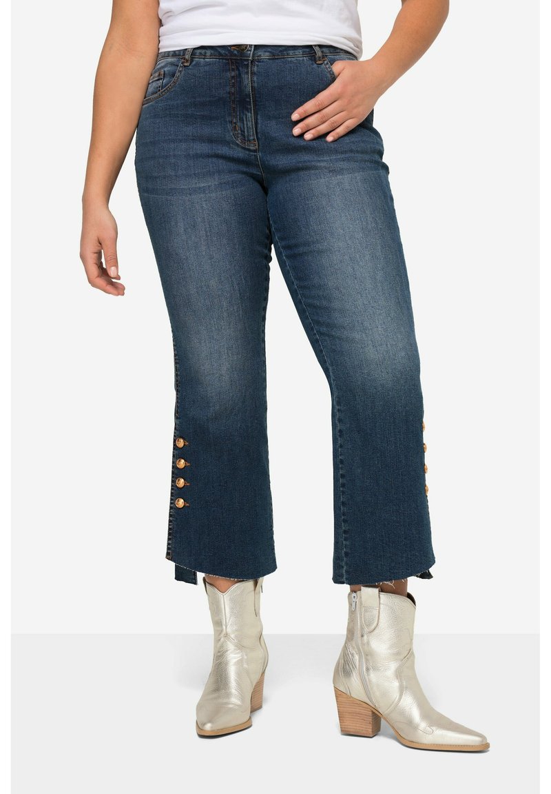 Jeans acortados de mezclilla oscura con dobladillo deshilachado y acentos de botones dorados a los lados, combinados con botines plateados metálicos.