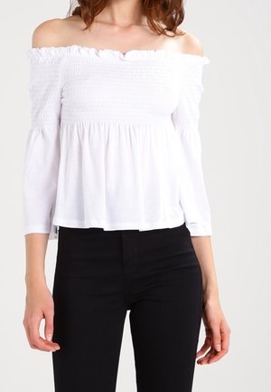 Kvinde iført en hvid off-shoulder langærmet top og sorte bukser med høj talje, står mod en ensfarvet baggrund.