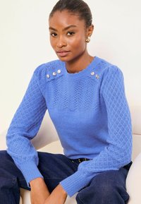 Jasnoniebieski dziergany sweter z fakturowanymi rękawami i wzorzystym korpusem. Zdobiony dekoracyjnymi guzikami na ramieniu i ściągaczami na mankietach.