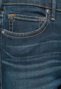 Tummansiniset farkut, joissa on pieni etutasku, vaaleanruskeat tikkaukset ja metallinen nappi vyötäröllä. Tasainen pinta.