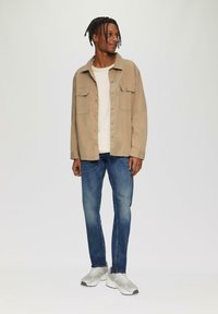 Beige Hemd mit Knöpfen und zwei Vordertaschen, getragen über einem cremfarbenen Pullover. Kombiniert mit blauen Jeans und hellgrauen Sneakers. Schlichtes Design.