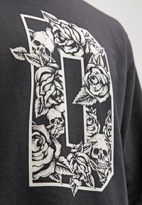 Tessuto nero con una grande lettera "D" bianca decorata con rose dettagliate e teschi intrecciati nella forma della lettera.