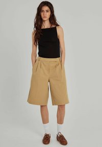 Svart åtsittande linne, beige veckade shorts, knä-långa, lätt tyg, bärs med vita strumpor och bruna snörskor. Neutral bakgrund.