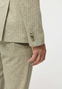 Férfi keze bézs textúrájú zakóujj mellett, négy gombbal és hozzáillő nadrággal, egyszínű háttér előtt.