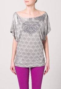 Top gris de corte suelto con diseño de hombros al descubierto, que presenta un patrón texturizado y acentos metálicos. Combinado con leggings morados brillantes.