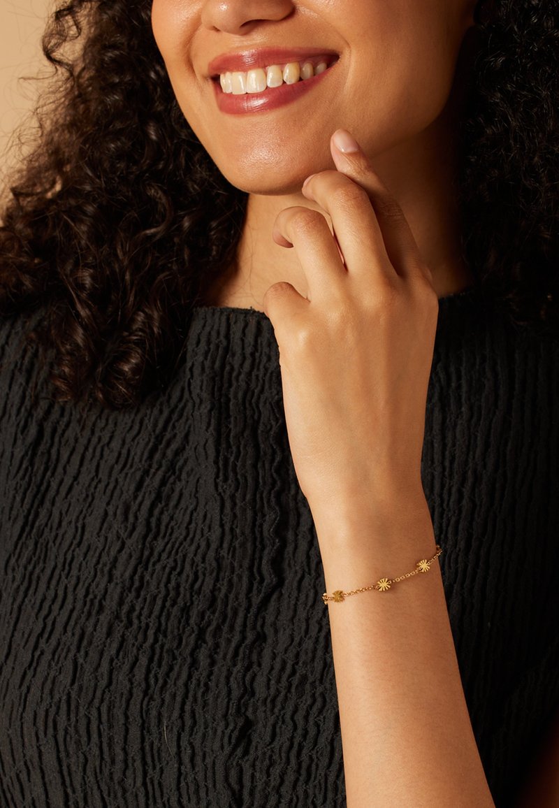 Gouden armband met bloemaccenten, fijn geschakeld ontwerp, gedragen over een textuur zwart kledingstuk. Huidskleur zichtbaar op de hand.