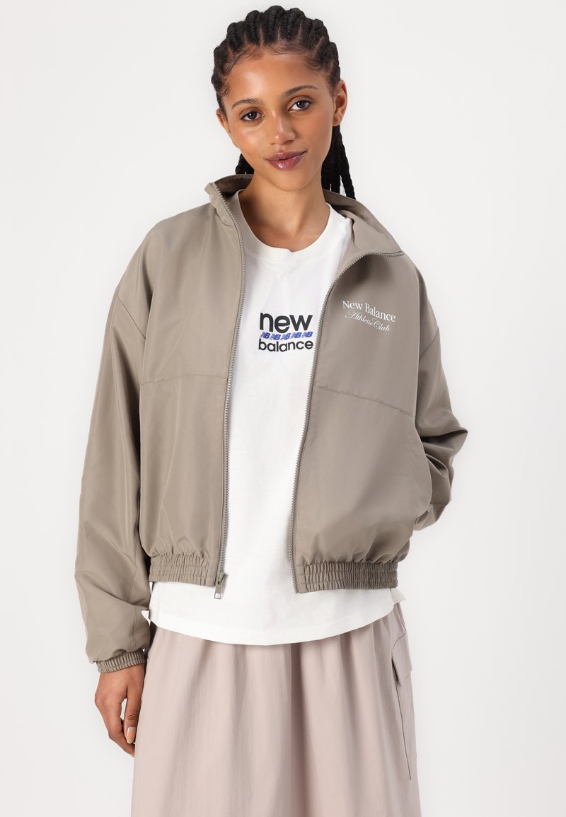 Blouson aviateur khaki avec fermeture éclair à l'avant, taille et poignets élastiques. T-shirt blanc dessous avec un logo en texte bleu et noir.