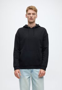 Crna majica s kapuljačom s džepom kangura, kapuljačom na vezice i rebrastim manšetama. Nošena s svijetloplavim trapericama koje imaju opušteni kroj.