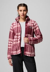 Giovane donna che indossa una giacca di pile a quadri maroon e rosa con tasca sul petto con cerniera, in piedi davanti a uno sfondo grigio uniforme.