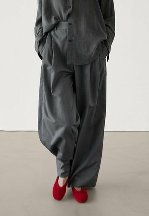 Osoba nosząca luźne ciemnoszare spodnie i pasującą zapinaną na guziki koszulę, ręce w kieszeniach, z jaskrawoczerwonymi butami stojąca na jednolitej podłodze.