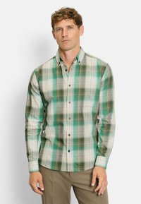 Mann mit hellbraunem, lockigem Haar, der ein kariertes Hemd in Grün und Beige sowie braune Hosen trägt und vor einem weißen Hintergrund steht.