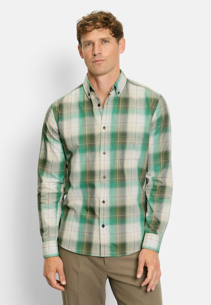 Mann mit hellbraunem, lockigem Haar, der ein kariertes Hemd in Grün und Beige sowie braune Hosen trägt und vor einem weißen Hintergrund steht.