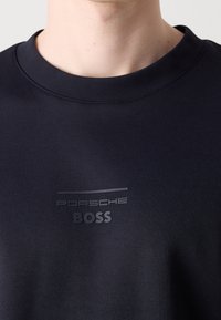 Gros plan sur une personne portant un t-shirt noir à col rond avec le texte embossé "Porsche BOSS" sur la poitrine.