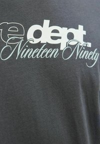 Grå t-shirt lavet af bomuld. Har stor hvid og lyseblå tekst, der står "e dept. Nineteen Ninety." Glat tekstur.
