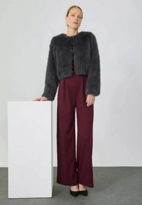 Grå faux fur kort jacka, ihopsatt med burgundy vida byxor. Modellen står bredvid en vit låda, iförd svarta spetsiga skor.