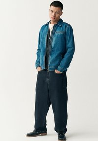 Chaqueta de mezclilla en azul claro con cierre delantero, dos bolsillos y cuello. Pantalones vaqueros de pierna ancha en azul oscuro. Zapatos negros con suelas marrones.