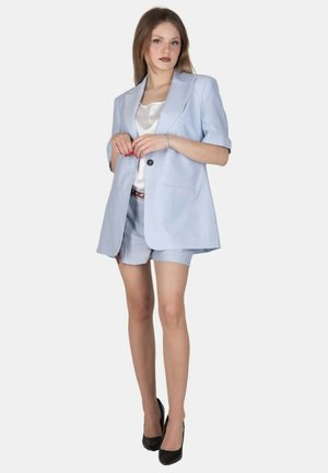 Giovane donna che indossa un blazer azzurro chiaro e pantaloncini abbinati con un top bianco in raso e tacchi alti neri, in piedi davanti a uno sfondo uniforme.