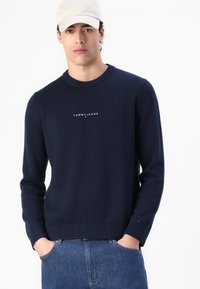 Tamno plava pletena majica s okruglim izrezom i rebrastim manžetama, s bijelim "TOMMY JEANS" logotipom na prsima. Meko na dodir.