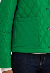 Grønn quiltet jakke med en firkantet lomme og svarte knapper, med en glatt tekstur og en tettsittende design, kombinert med et kremfarget innerlag.