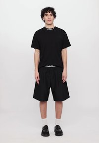 Melna t-krekla ar apaļu kakla izgriezumu un mazu kabatu, kopā ar brīvām melnām šortiem. Apavi ir melni ar gludu apdari. Redzami balti zeķes.