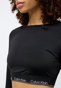 Μαύρο μακρυμάνικο τοπ από λείο ύφασμα, με εφαρμοστή σχεδίαση και λωρίδα με το λογότυπο Calvin Klein στο τελείωμα.